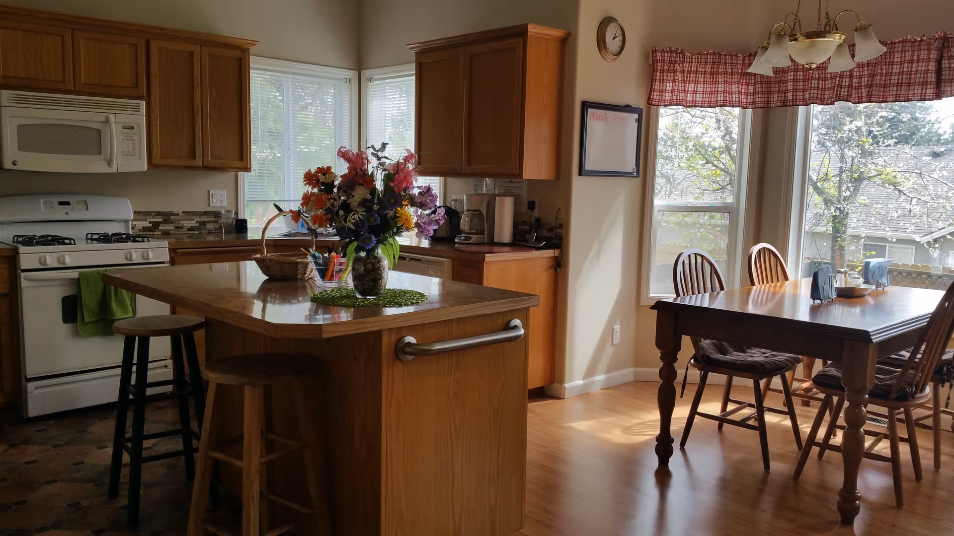 A bright kitchen and dining area with wooden cabinets and a white stove and microwave. A kitchen island with a granite countertop has a vase of colorful flowers and two wooden stools. The dining area features a wooden table with four chairs, positioned near large windows with red checkered valances, letting in natural light and showing a view of trees outside.