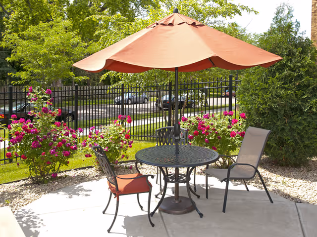 Outdoor patio area with a round metal table and two chairs, one with a red cushion and the other without, shaded by a large red umbrella. Surrounding the patio are green bushes and vibrant pink flowers, with a black metal fence and parked cars visible in the background.