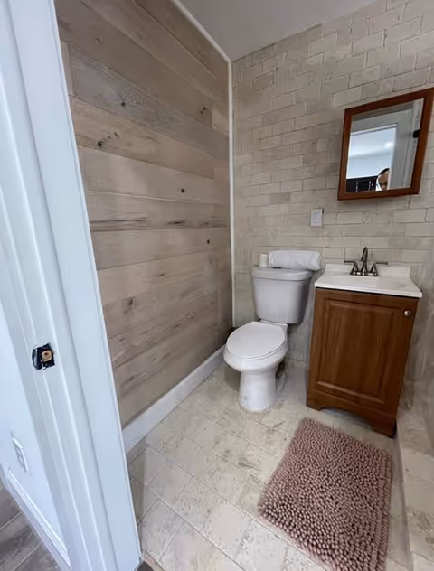 Small bathroom with a toilet, wooden vanity and mirror, tiled walls and a wood-paneled accent wall, and a pink bath mat on the floor.
