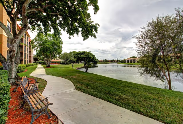 A curved concrete walkway beside a calm lake with green grass and trees on both sides. There are two wooden benches with metal armrests along the path. On the left side, a multi-story building with balconies is partially visible. The sky is cloudy.