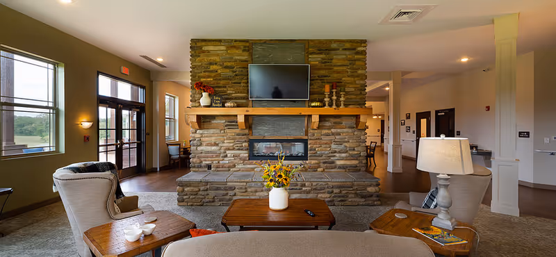 A cozy communal living room featuring a stone fireplace with a mounted TV, comfortable armchairs and tables, and a vase of flowers on a central coffee table.