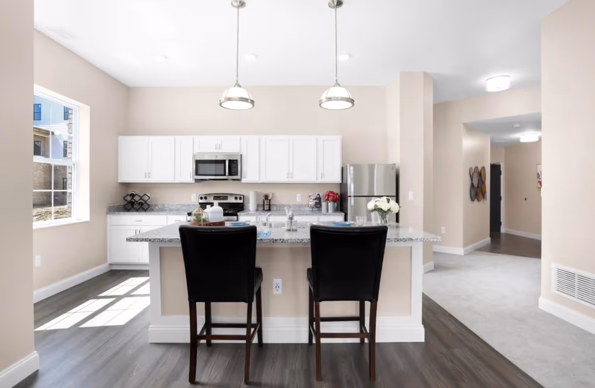 Modern kitchen with white cabinets, stainless steel appliances including a microwave, stove, and refrigerator. A granite countertop island with two black chairs is in the foreground, with pendant lights hanging above. There is a window on the left letting in natural light, and a hallway visible to the right.