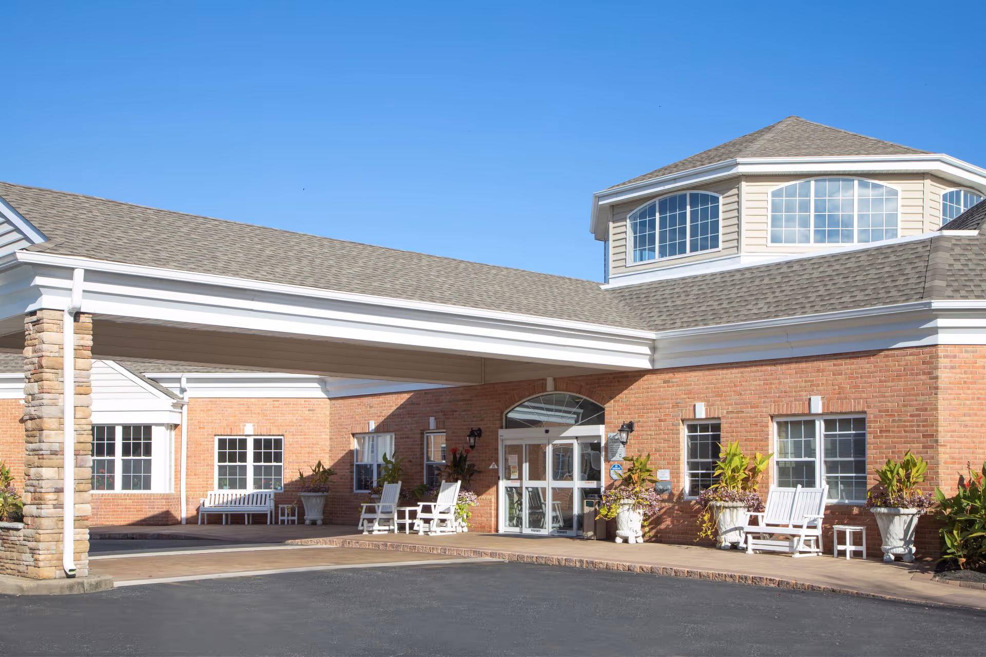 Exterior view of Pines Village facility entrance with a covered drop-off area, brick walls, large windows, white benches, potted plants, and a clear blue sky.