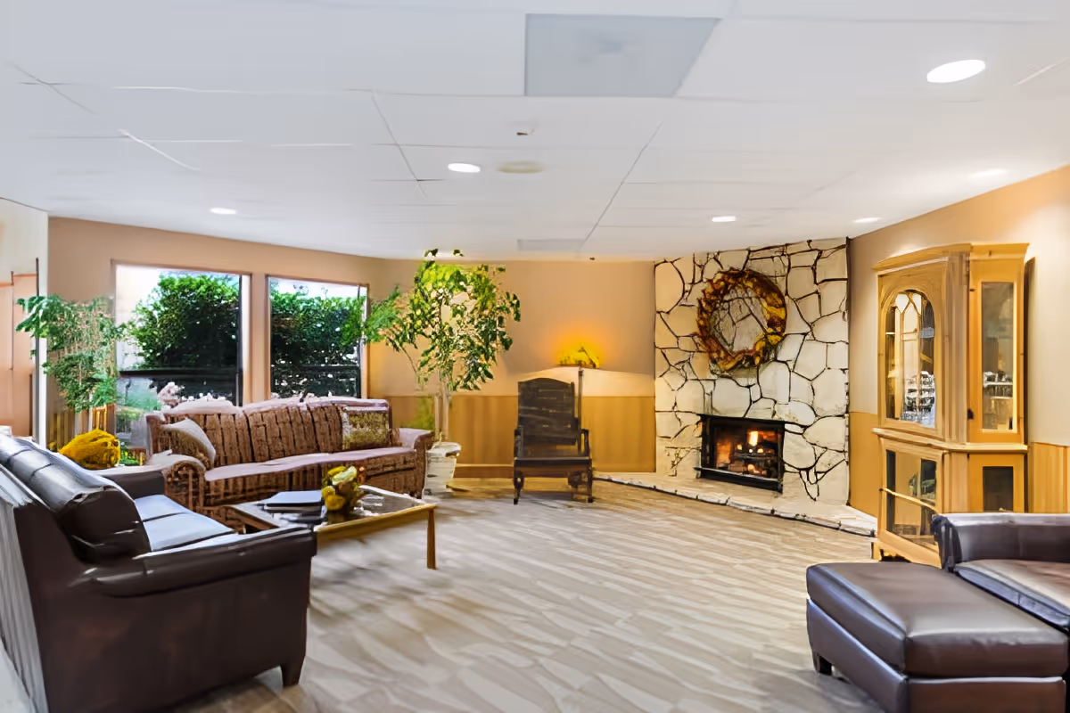 A cozy living room in a retirement and assisted living facility featuring a stone fireplace with a wreath above it, a wooden rocking chair, a glass-front wooden cabinet, a brown leather sofa with matching ottoman, a wicker sofa, a glass coffee table with decorative items, and several green plants near large windows.