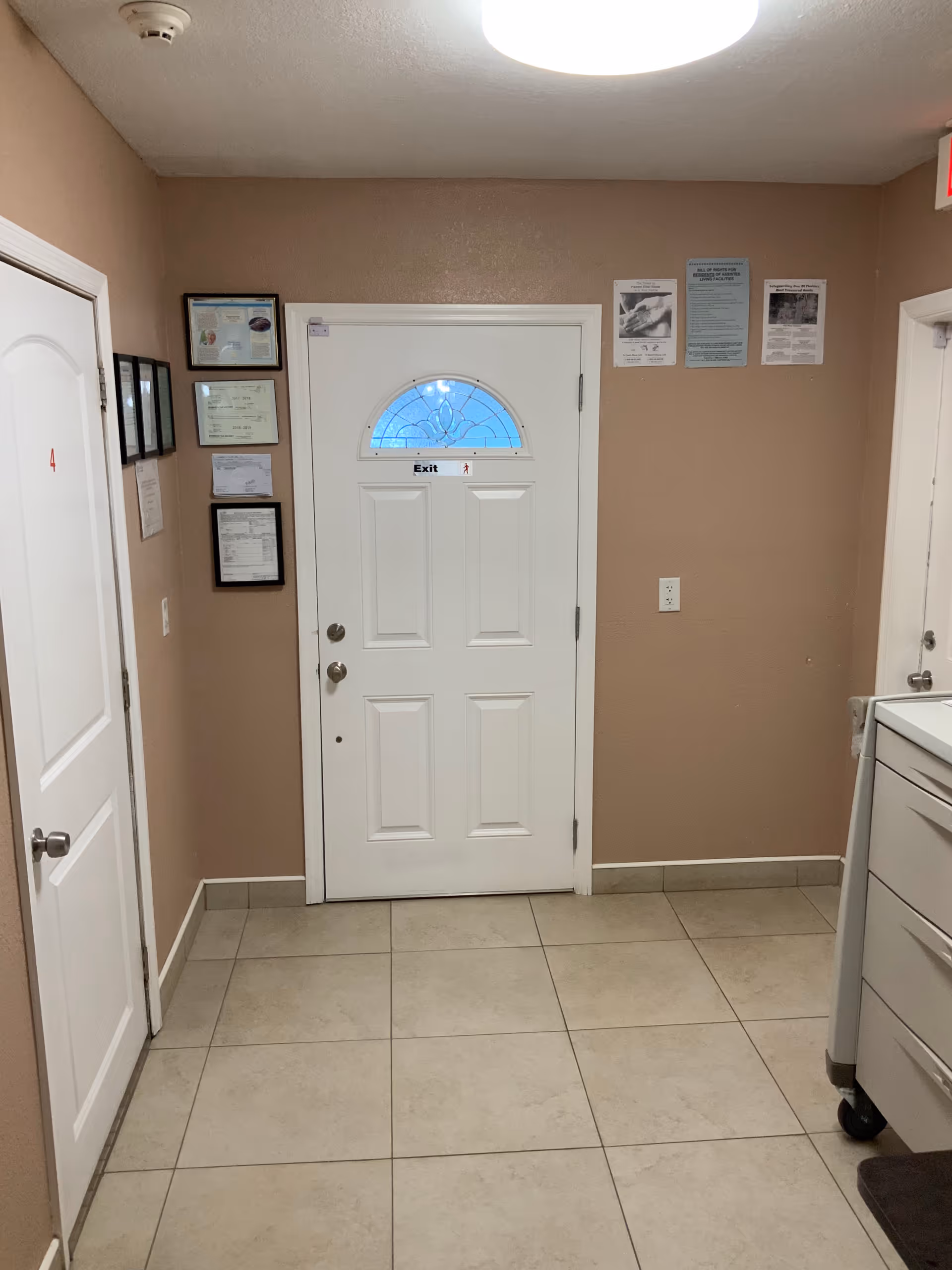 Interior view of a hallway in a senior living facility with beige walls and tiled floor. There is a white door with a small decorative window and an 'Exit' sign above the handle. To the left, there is another white door labeled with the number 4 and several framed certificates or notices on the wall. On the right side, part of a white cabinet or cart is visible. Several informational posters are displayed on the wall above the exit door.