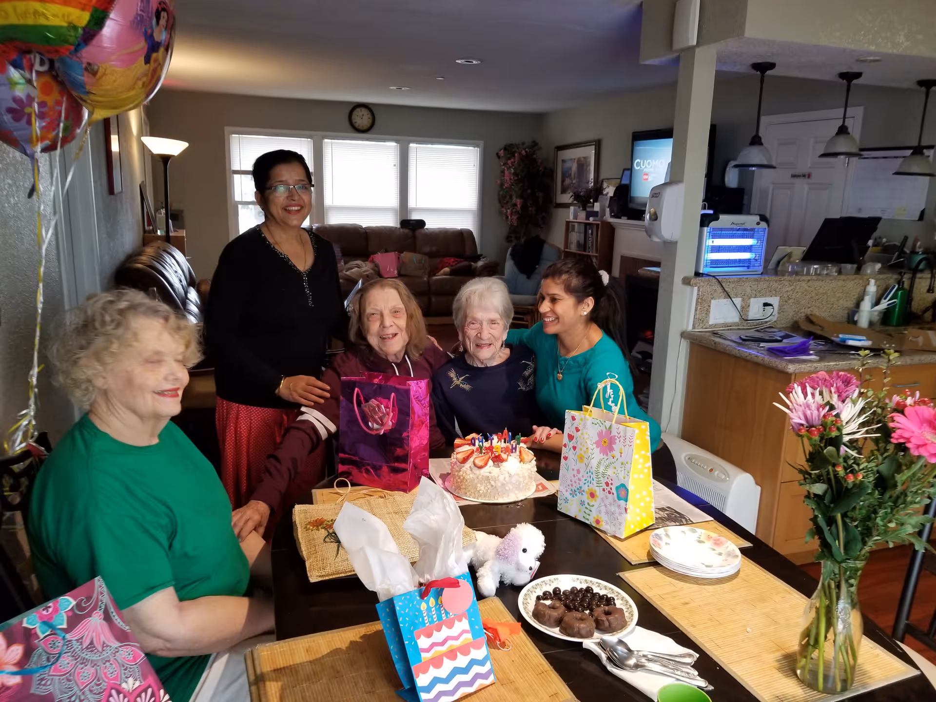 A group of five women gathered around a dining table celebrating a birthday. There is a birthday cake with lit candles on the table, along with gift bags, a small stuffed dog, and plates with chocolate treats. The setting is a cozy residential living space with a kitchen visible in the background. Balloons are seen on the left side of the image.