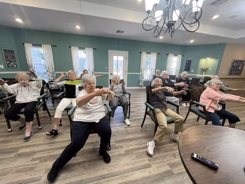 A group of elderly individuals seated on chairs in a spacious room with wooden flooring and teal walls, participating in a seated exercise session with arms extended forward. The room has multiple windows with white curtains, a chandelier hanging from the ceiling, and a round table with a remote control in the foreground.