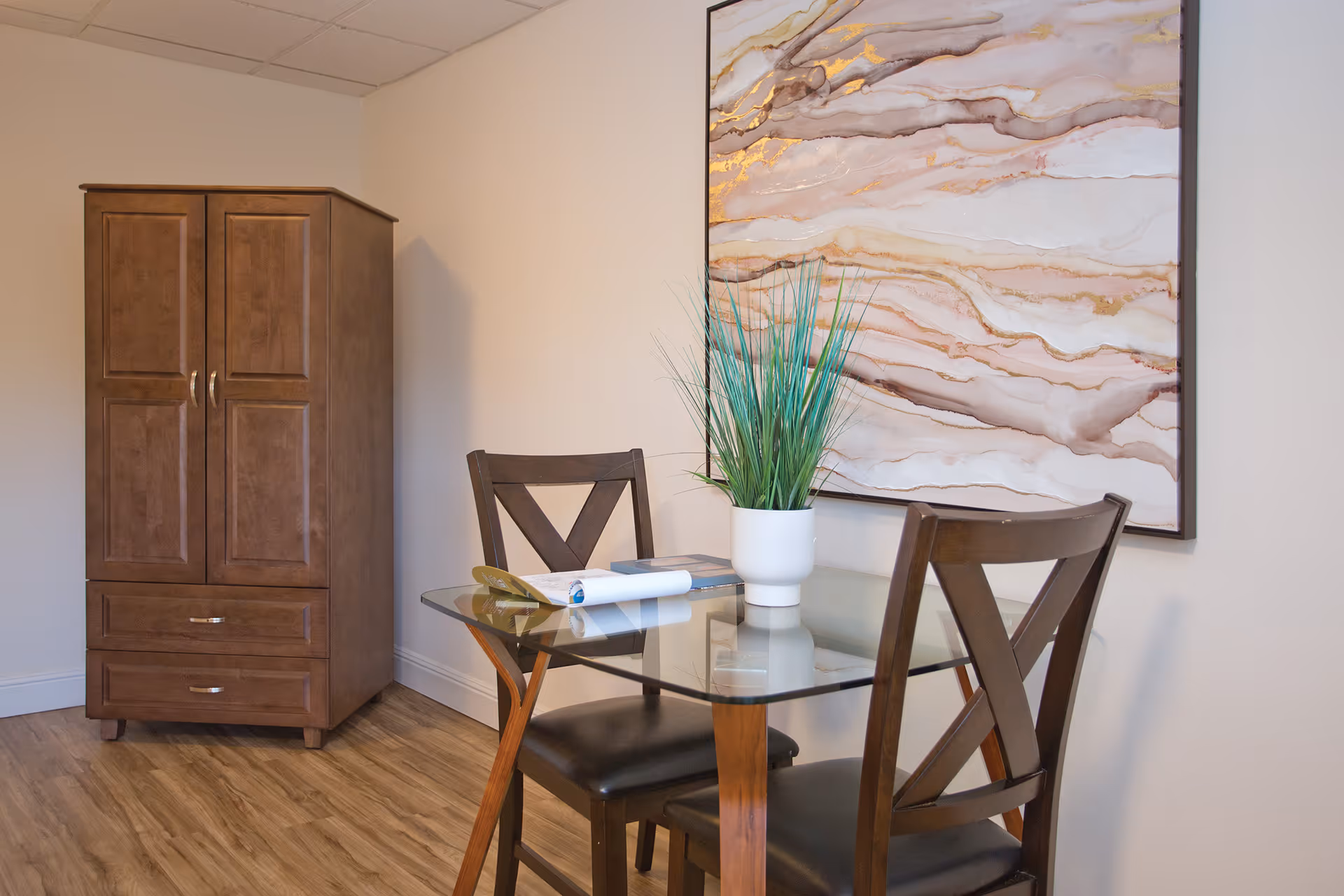 A small dining area with a glass-top table and two wooden chairs with black cushions. On the table is a white pot with green artificial grass and some reading materials. Behind the table is a large abstract wall art with shades of beige, white, and gold. To the left is a wooden cabinet with two doors and two drawers. The floor is wood laminate and the walls are light-colored.