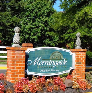 A brick sign at the entrance of Morningside Assisted Living, displaying the facility name and address 2800 Polo Parkway, surrounded by flowers and greenery with trees in the background.