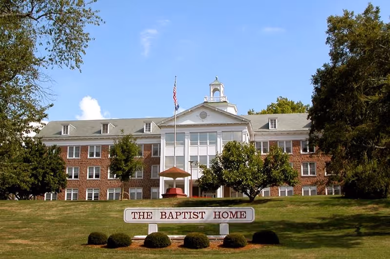 Front exterior view of a large brick building with white columns and a cupola on the roof, surrounded by trees and a well-maintained lawn. A sign in front reads 'THE BAPTIST HOME' and an American flag flies on a flagpole near the entrance.