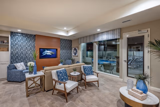 A modern and cozy living room area in a senior living facility featuring a beige sofa, two white cushioned chairs with blue patterned pillows, a blue armchair, and a wall-mounted TV displaying an image of a shark. The room has a tiled floor, decorative plants, and large windows looking out to an outdoor pool area with lounge chairs.