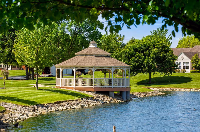 A white wooden gazebo sits at the edge of a pond surrounded by green lawn, trees, and nearby residential buildings.