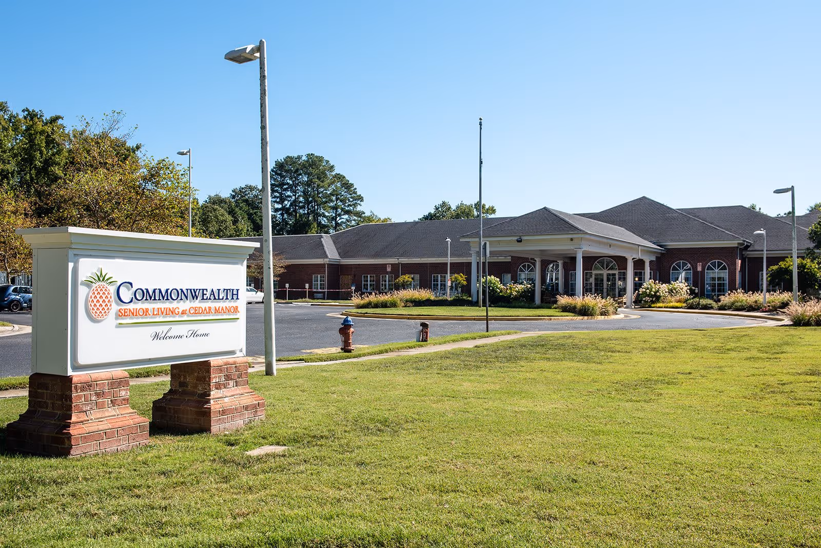 Exterior view of Commonwealth Senior Living at Cedar Manor facility with a large sign in the foreground displaying the facility name and a well-maintained lawn leading up to the building under a clear blue sky.
