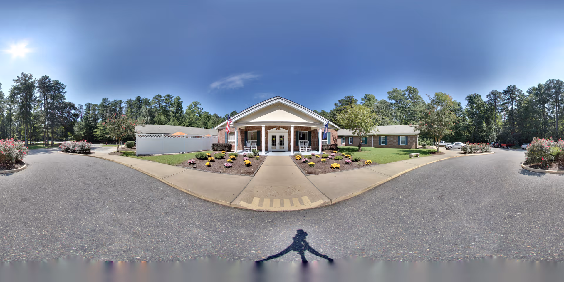 Front entrance of a single-story senior living building with a covered portico, flags, flowerbeds and a circular driveway surrounded by trees.
