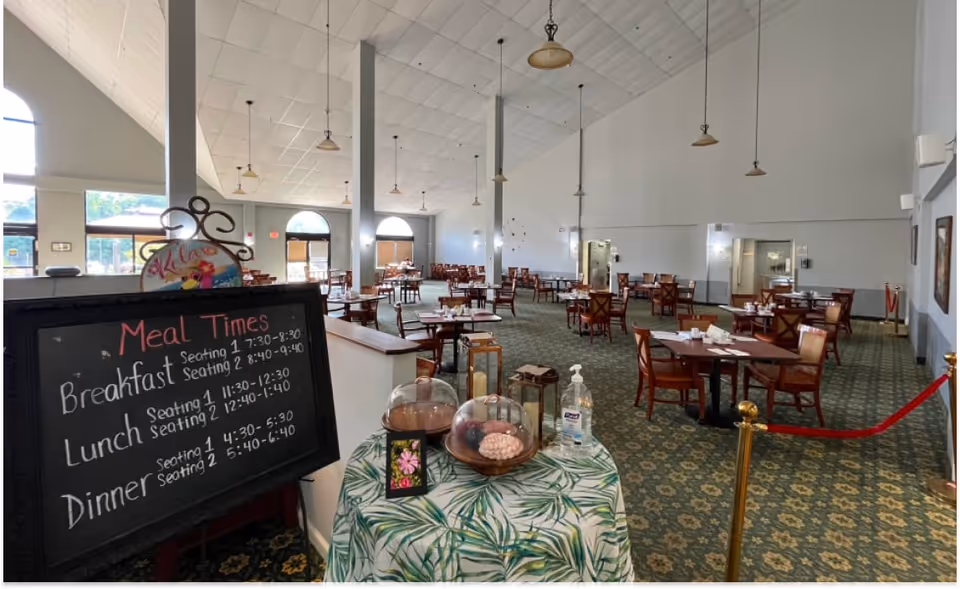 Large dining room with many tables and chairs, a chalkboard listing meal times, and a small front table with hand sanitizer.