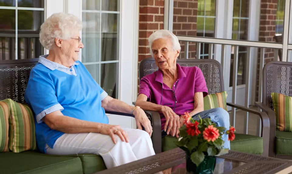Two elderly women sitting and chatting on a patio with green cushioned chairs and a glass table with a potted plant with red flowers in front of them. The background shows large windows and a brick wall.