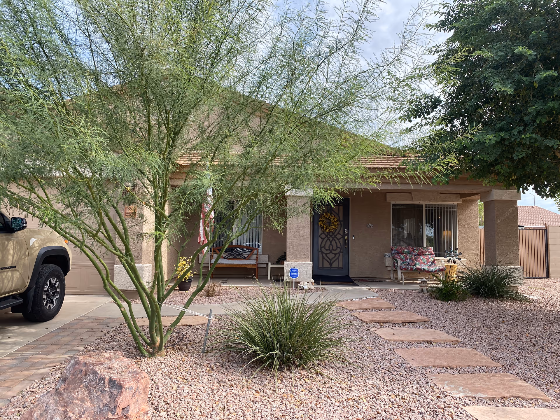 Front exterior view of a single-story house with a covered porch, two chairs with floral cushions, a decorative wreath on the front door, and a pathway made of stepping stones leading through a gravel yard with desert plants and trees. A beige vehicle is partially visible parked on the driveway to the left.