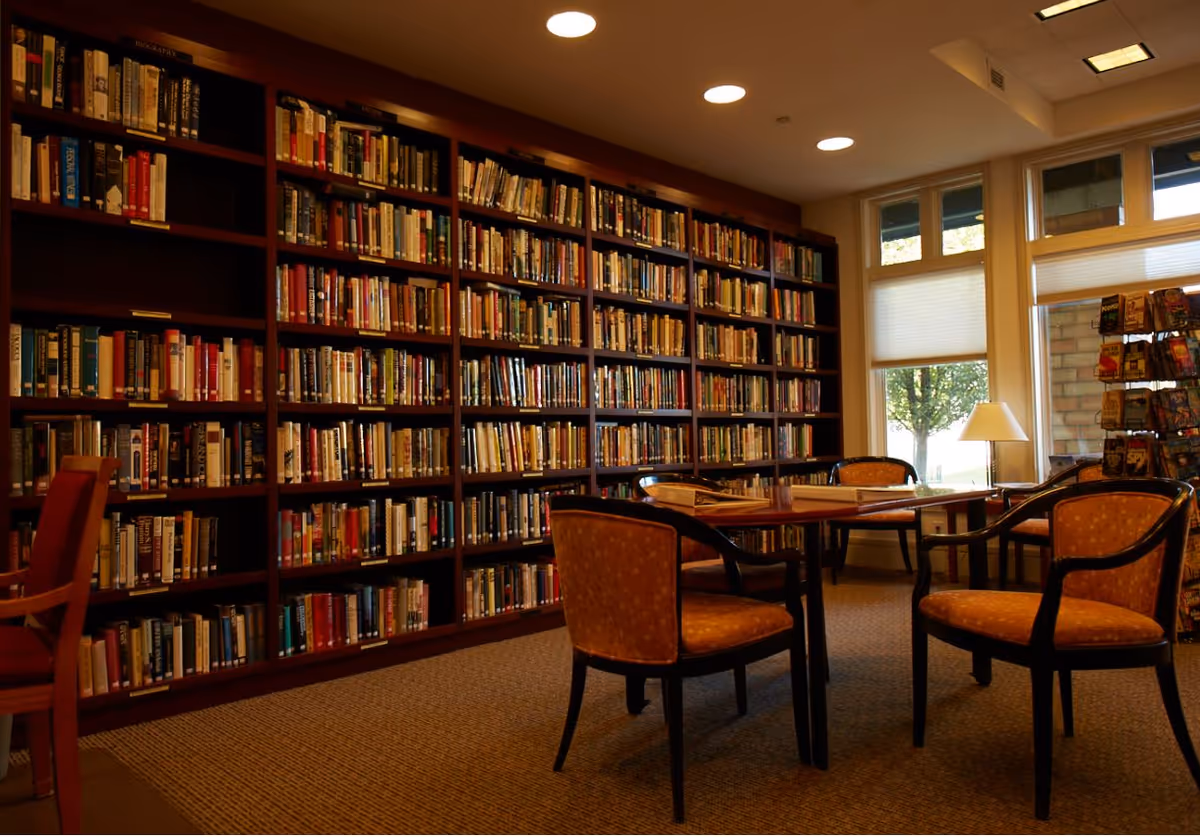 A cozy library room with tall wooden bookshelves filled with books along one wall. There are several cushioned chairs around a wooden table in the center of the room. Large windows let in natural light, and a small lamp is placed on a side table near the windows.