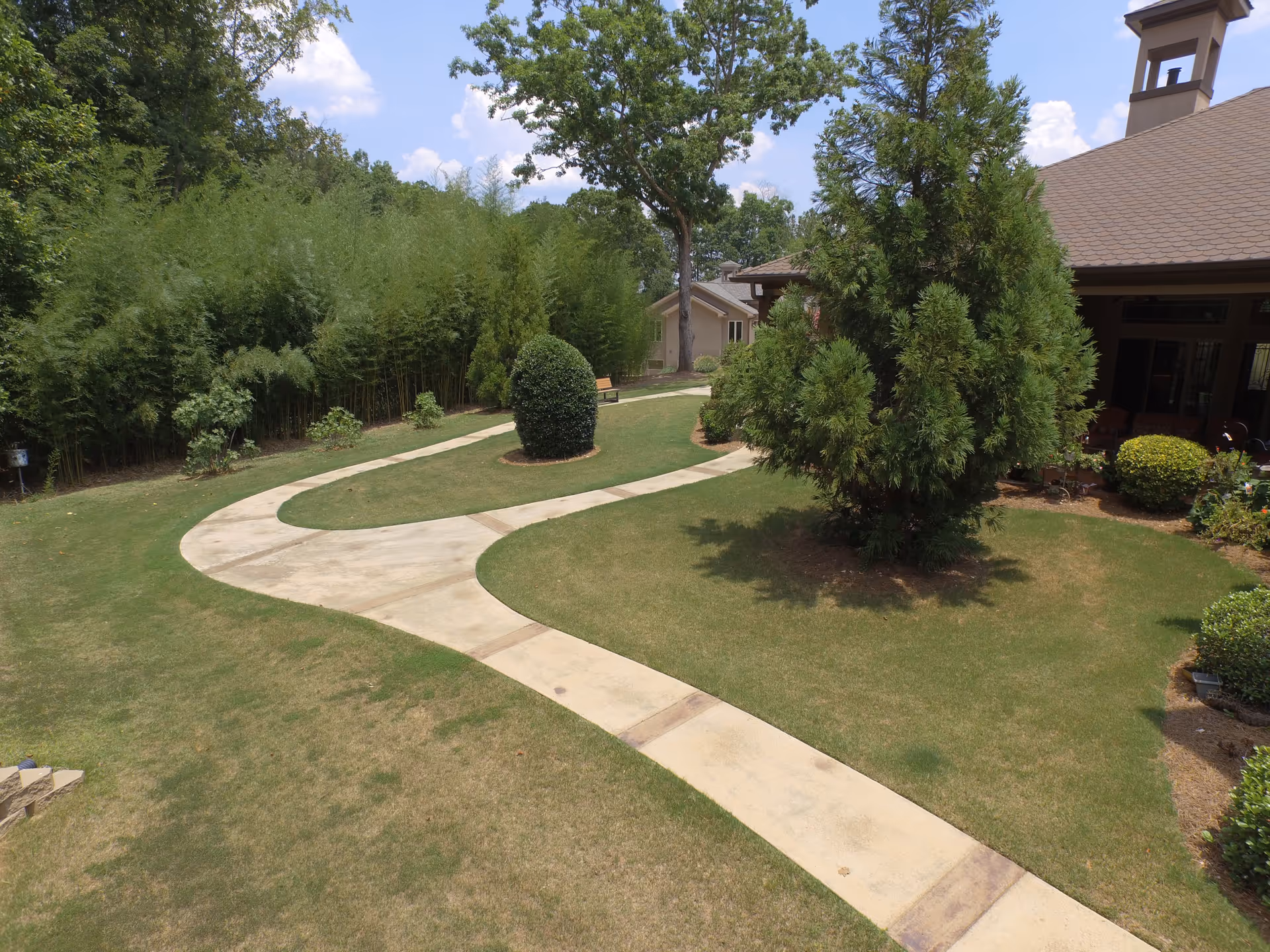 Curved concrete walking paths through a green lawn with various trees and shrubs, adjacent to a building with a brown roof and a bell tower under a partly cloudy sky.