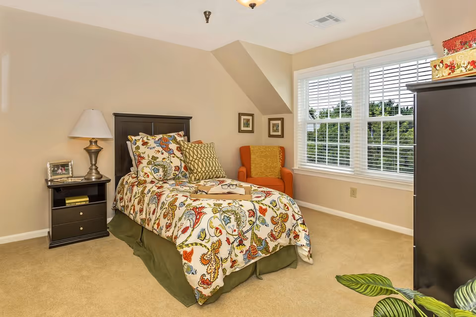 Bright bedroom featuring a single bed with colorful floral bedding, a nightstand and lamp, an orange armchair by a large window, and a dark dresser.