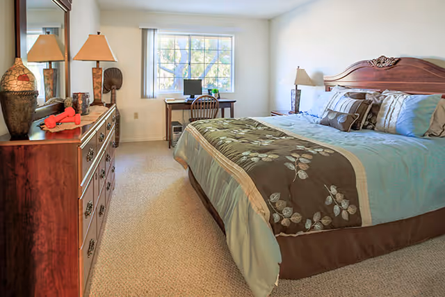 A well-lit bedroom with a large bed featuring a decorative wooden headboard and a floral-patterned comforter. To the left, there is a wooden dresser with a mirror above it, adorned with a lamp, decorative items, and a small red object. At the far end of the room, a window lets in natural light, and beneath it is a small desk with a chair and a computer monitor.
