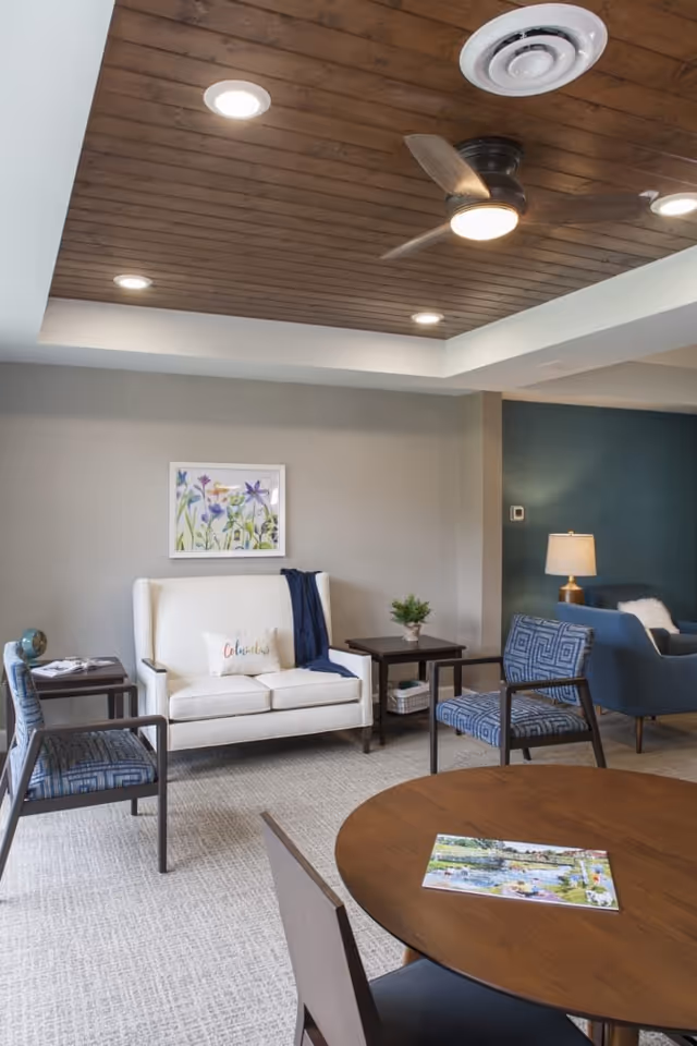A cozy living room area with a white loveseat adorned with a decorative pillow and a dark blue throw blanket. Two patterned blue armchairs and a wooden round table with a magazine on it are also visible. The room features a wooden ceiling with recessed lighting and a ceiling fan, a framed floral artwork on the wall, and a side table with a small plant and a lamp in the background.