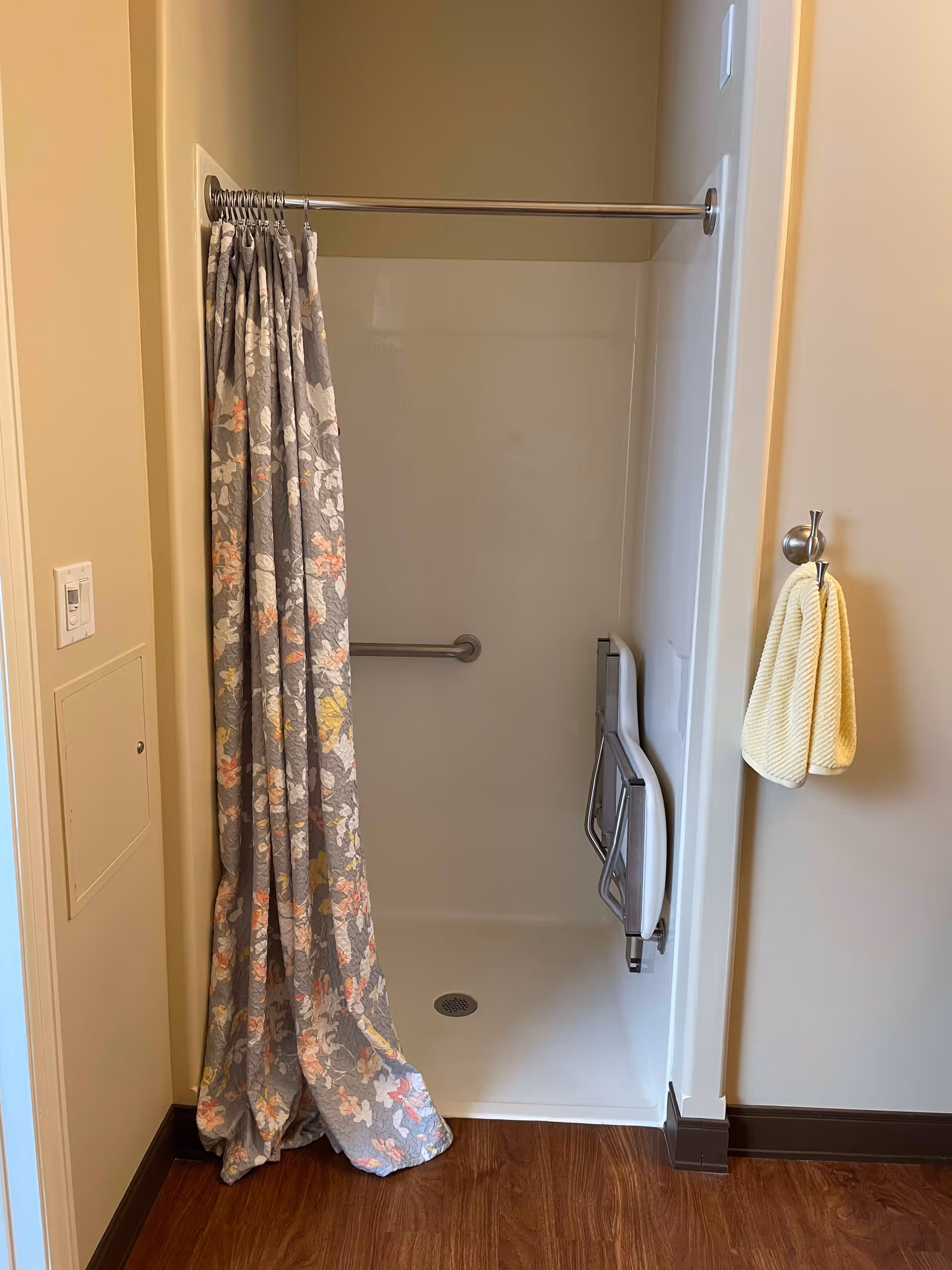 A small shower stall with a floral-patterned shower curtain partially drawn to the side. Inside the shower, there is a foldable white shower seat attached to the wall and a horizontal grab bar. A yellow hand towel hangs on a hook on the wall outside the shower. The floor outside the shower is wood, and the walls are painted beige.