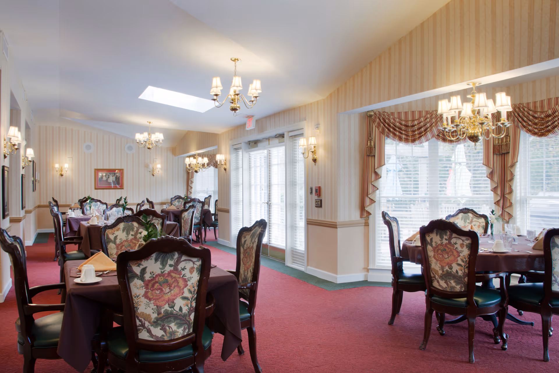 Formal dining room with multiple set tables, floral-upholstered chairs, chandeliers, and large windows with drapes.