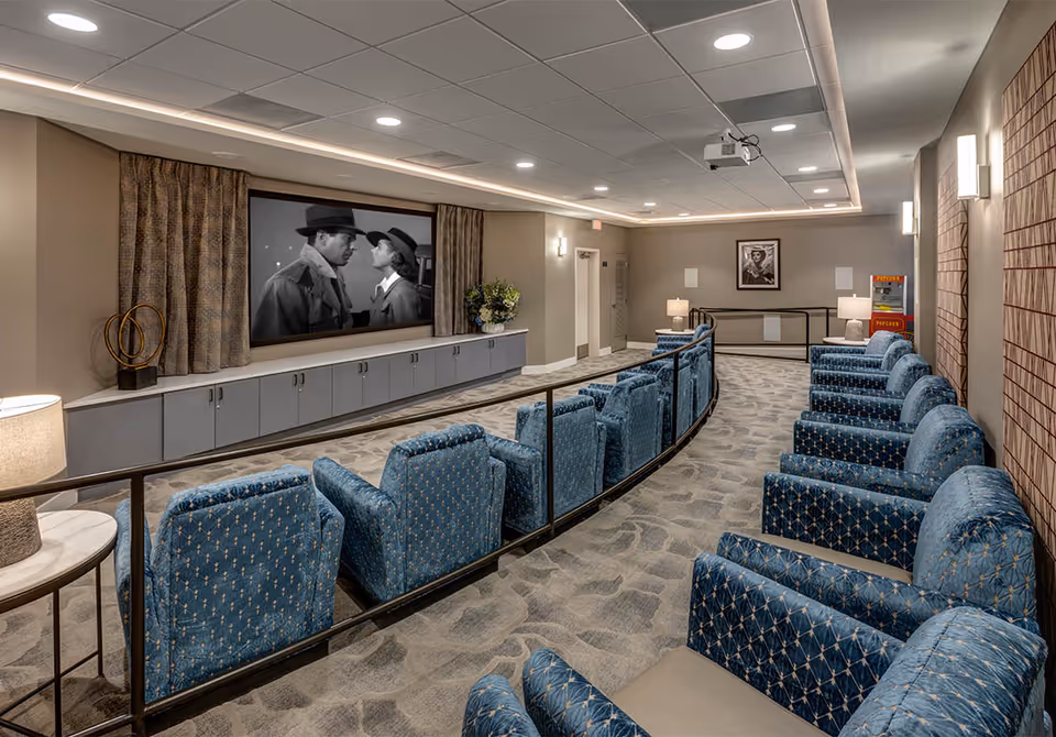 A senior living facility's private theater room with rows of blue upholstered seats facing a large screen showing a black-and-white movie.