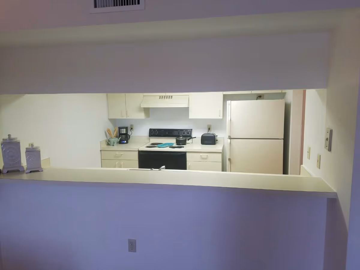 Pass-through view into a compact kitchen with white cabinets, an electric stove, refrigerator, toaster and coffee maker.