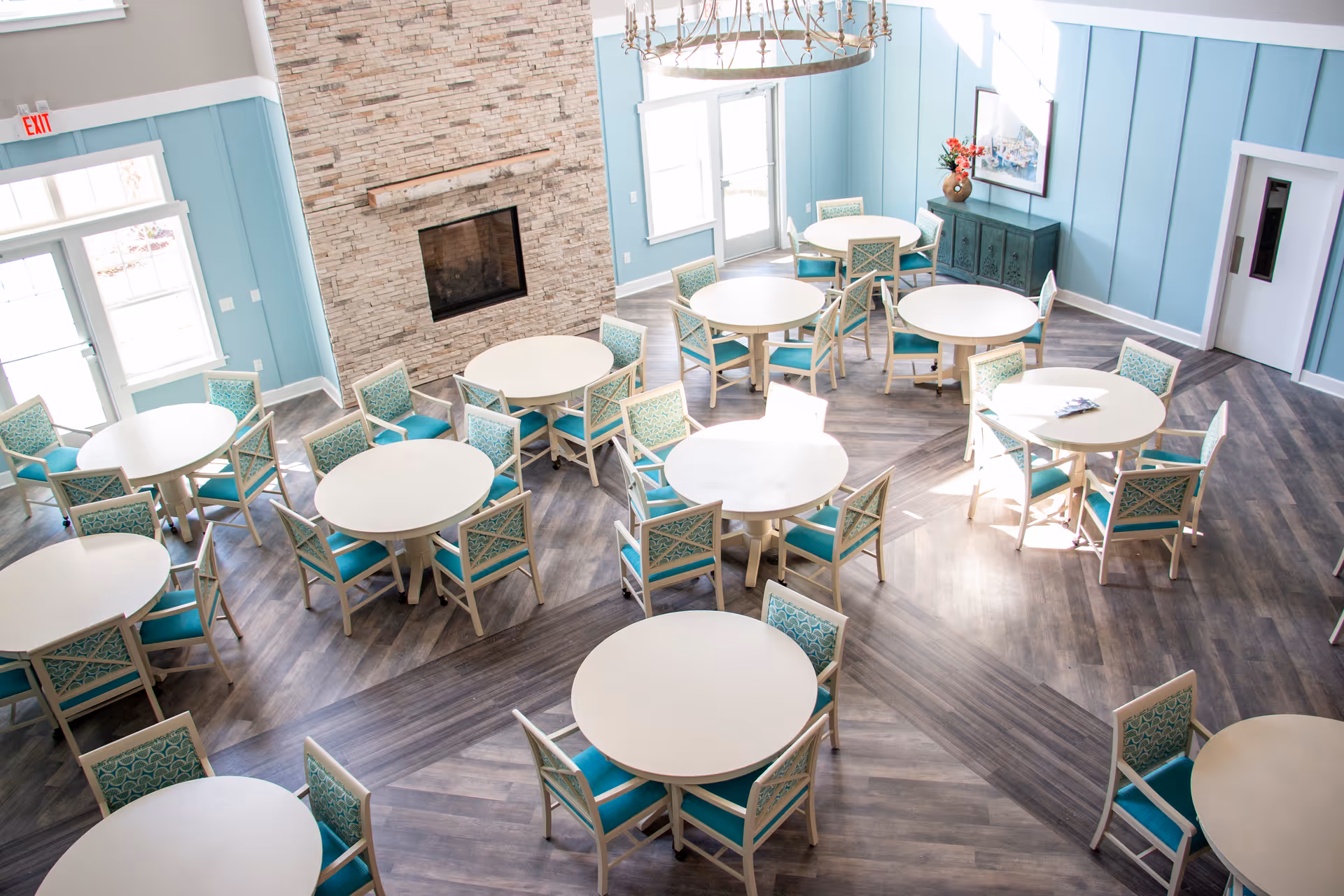 Bright communal dining/activity room with multiple round tables and teal-upholstered chairs arranged on wood-look flooring in front of a stone fireplace.