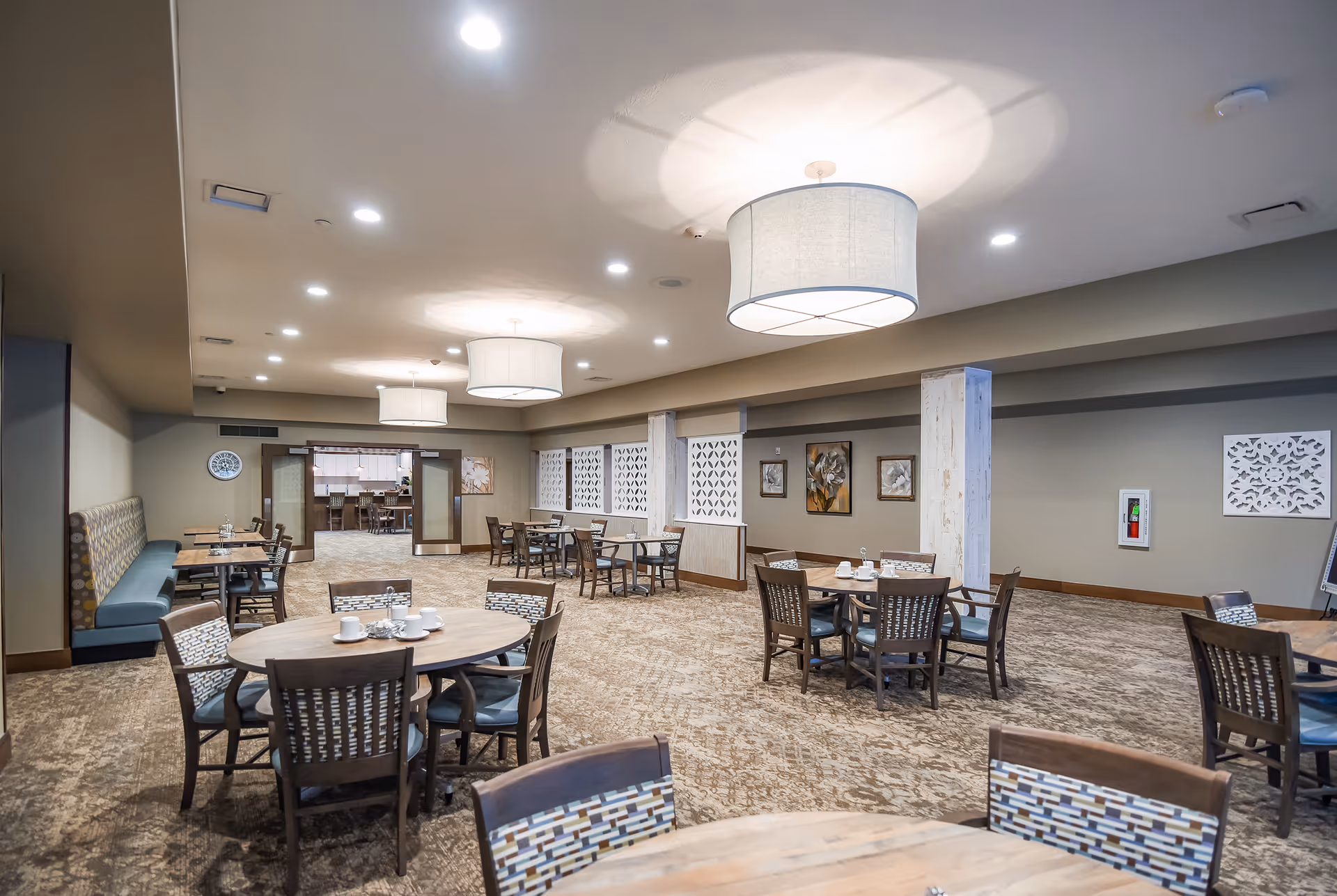 A spacious dining room with multiple round and rectangular tables surrounded by chairs. The room features patterned carpet, modern pendant lights hanging from the ceiling, and decorative wall art. There is a seating bench along one wall and a doorway leading to another room in the background.