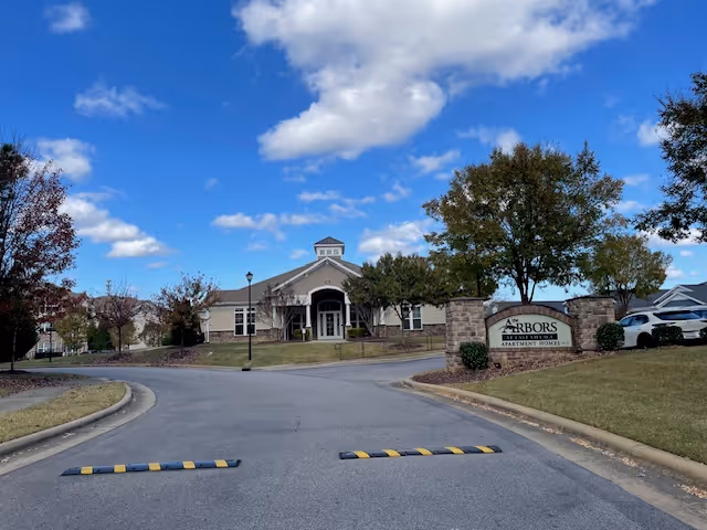 View of the entrance to a senior living facility named The Arbors at East Village, showing a single-story building with a peaked roof, surrounded by trees and a curved driveway with speed bumps under a partly cloudy blue sky.