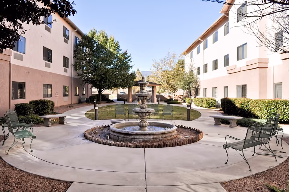 Sunny courtyard between residential buildings with a central tiered fountain, benches, and paved walkways.