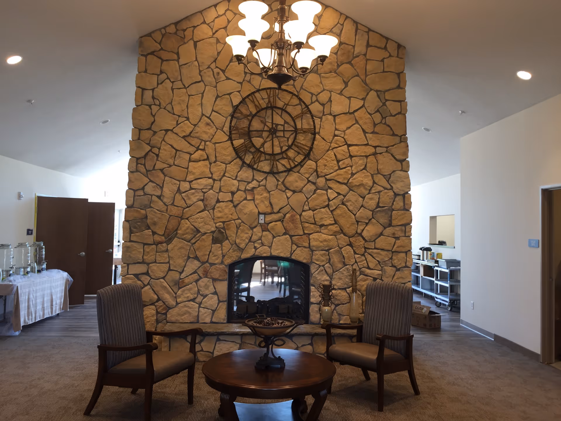 Interior of a senior living facility featuring a stone fireplace with a large decorative clock above it. Two upholstered armchairs are positioned on either side of a round wooden coffee table in front of the fireplace. A chandelier with multiple lights hangs from the ceiling above. The room has carpeted flooring and light-colored walls, with open doorways and tables visible in the background.