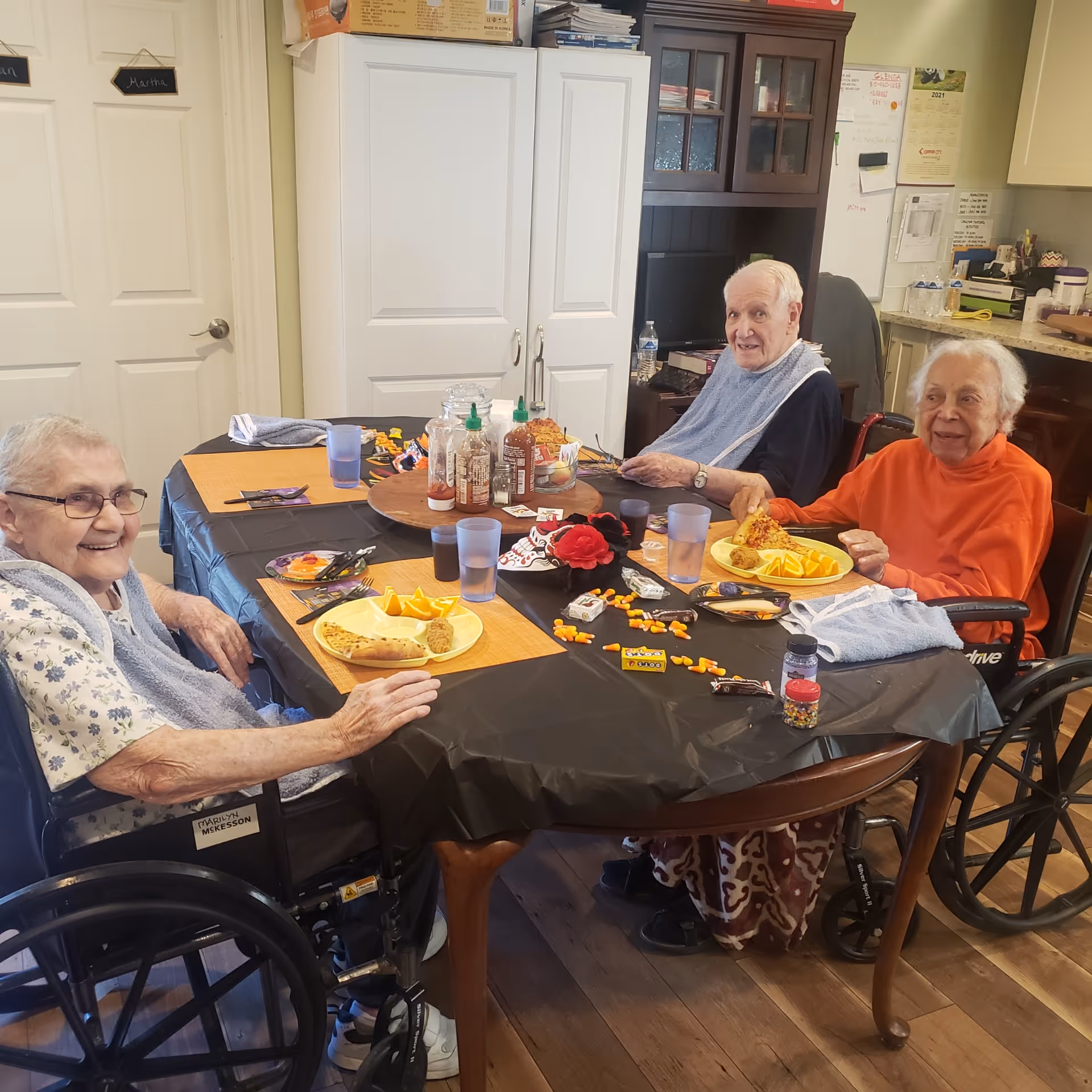 Three elderly individuals sitting around a dining table in a residential care facility, enjoying a meal with plates of food including pizza and orange slices. The table is covered with a black tablecloth and has condiments and snacks on it. Two of the individuals are in wheelchairs, and the room has cabinets and a kitchen area in the background.