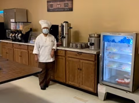 A masked chef in a white uniform stands in a kitchenette/serving area with wooden cabinets, beverage dispensers, and a small refrigerated display case.