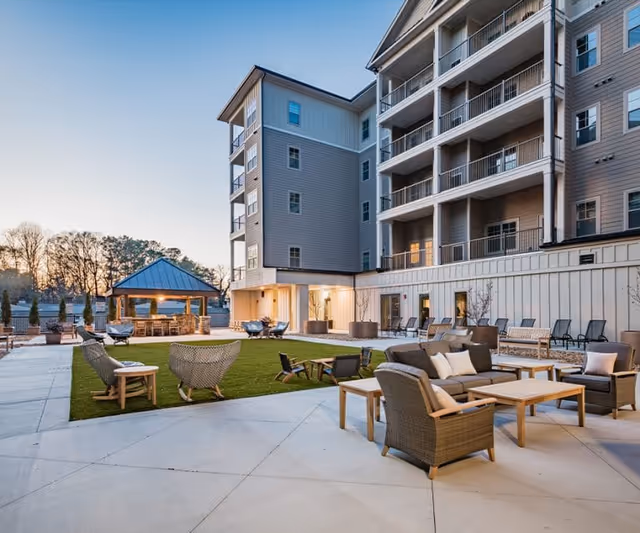 Outdoor patio area of a senior living facility with comfortable wicker seating, tables, and a small grassy area. The multi-story building with balconies overlooks the patio, and there is a covered pavilion in the background. The scene is captured at dusk with soft lighting.