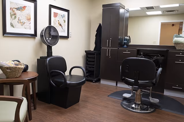 Interior of a salon area in a senior living facility with two black salon chairs, a hair dryer chair, a wooden side table with a basket, framed bird artwork on the wall, and dark wood cabinetry with a large mirror.