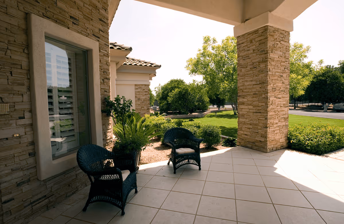 A covered outdoor patio area with two black wicker chairs with cushions, a potted plant, stone pillars, and a view of green bushes and trees in a landscaped garden.