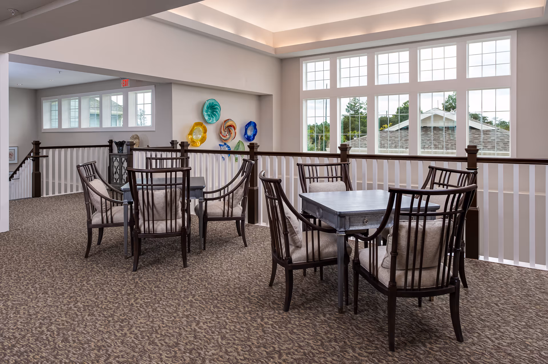 A bright and spacious interior seating area with two small square tables, each surrounded by four cushioned wooden chairs. The space features large windows letting in natural light, a carpeted floor, and decorative colorful glass wall art. A railing separates the seating area from a lower level.
