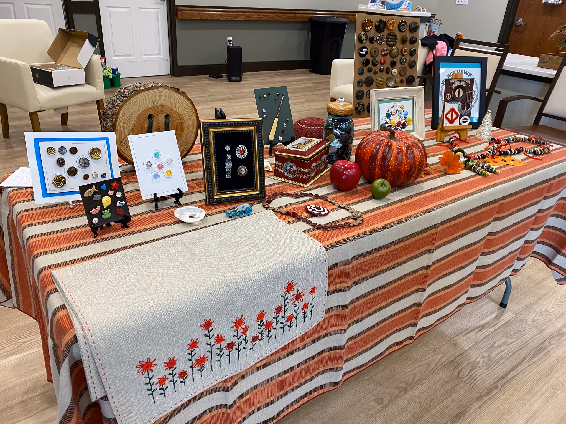 A table covered with a striped tablecloth and a beige runner with red flower embroidery, displaying various decorative items including framed button art, a wooden log slice, a pumpkin, artificial apples, a jar, a decorative box, and beaded necklaces in a room with wooden flooring and chairs in the background.