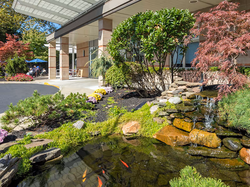 Front entrance of a senior living facility featuring a covered porte-cochère, landscaped pond with a small waterfall and koi, and outdoor seating.