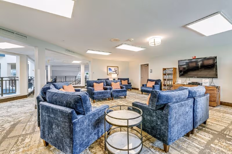 A spacious senior living common area with multiple blue upholstered armchairs arranged in a circle around a small round table. The room has a patterned carpet, white walls, and ceiling lights. A flat-screen TV is mounted on the wall with a wooden media console below it, and a bookshelf is nearby. The area overlooks an open space with railings and windows.