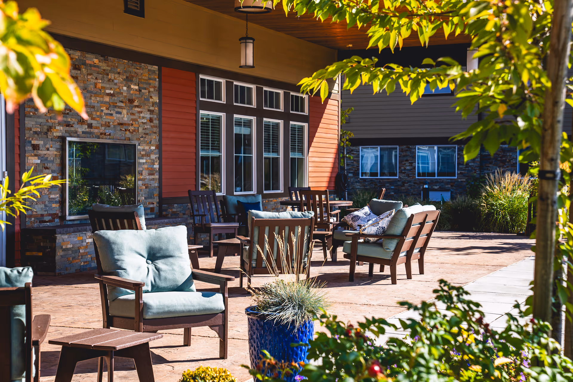 Outdoor patio area at Fieldstone on Clear Creek with cushioned wooden chairs and small tables arranged on a stone-paved surface. The patio is adjacent to a building with large windows and a stone and red siding exterior. There are plants and greenery around the patio, with a hanging lantern light above.