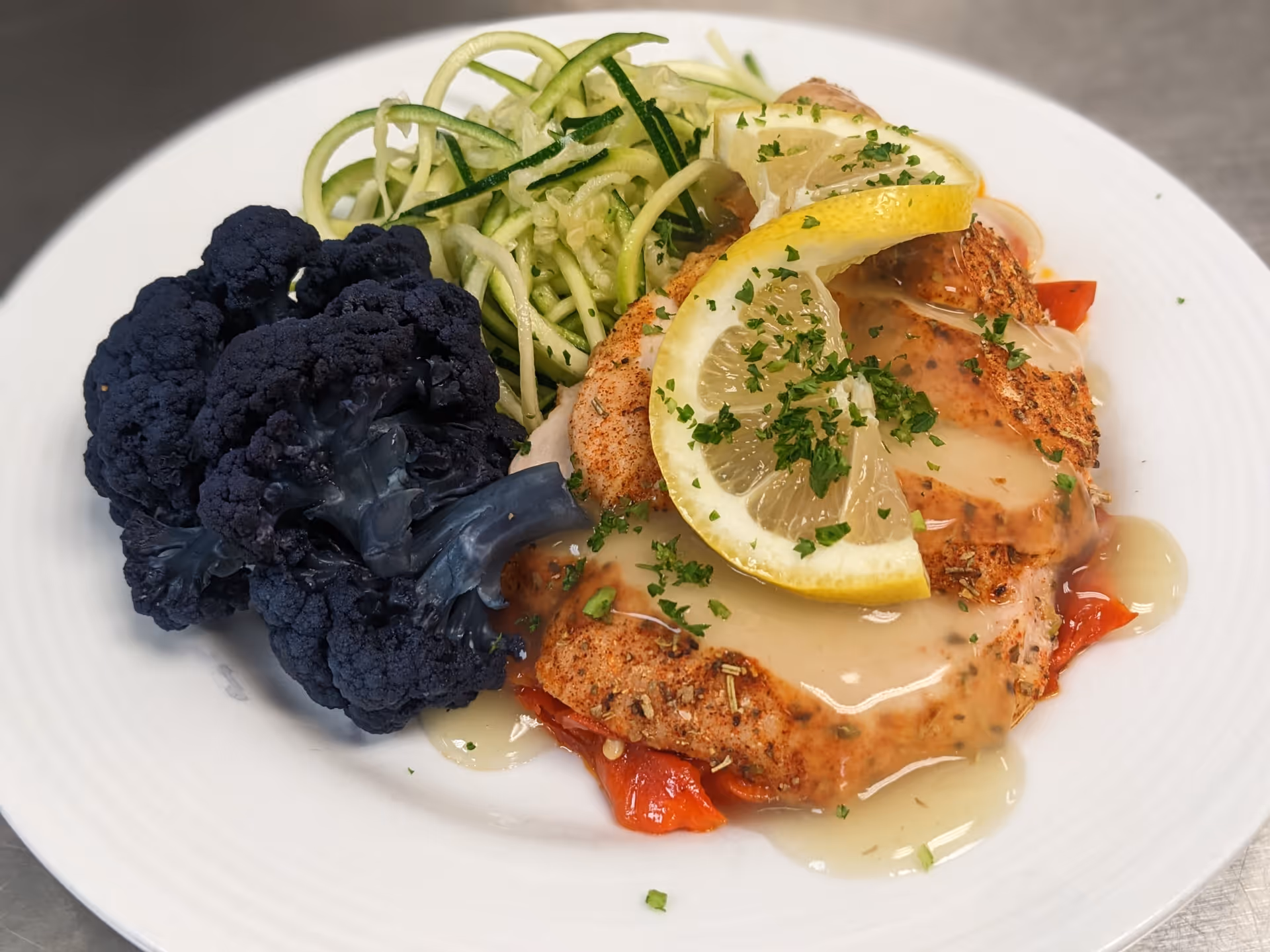 A white plate with a serving of seasoned cooked chicken breast topped with lemon slices and a light sauce, accompanied by dark purple cauliflower florets and spiralized zucchini noodles.
