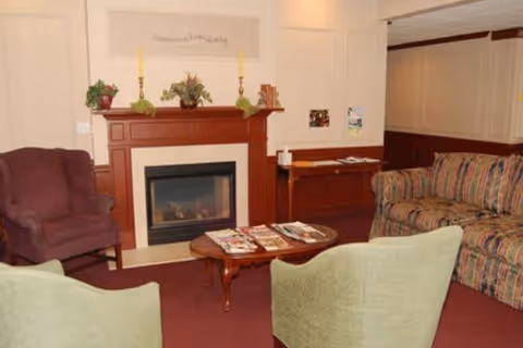 A cozy living room area with a wooden fireplace mantel decorated with plants, candles, and books. There are four chairs arranged around a wooden coffee table with magazines on it. The chairs include a purple armchair, a striped sofa, and two green armchairs. The walls are light-colored with wood paneling on the lower half.