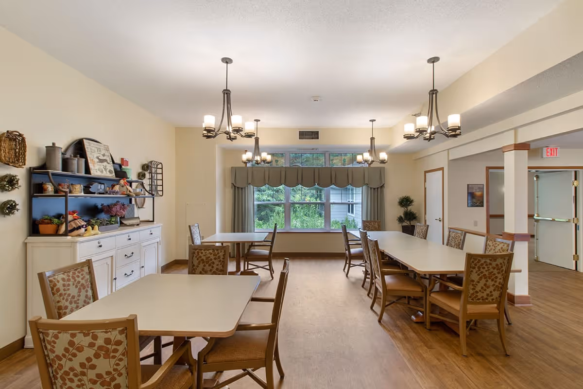 A well-lit dining room with multiple tables and chairs arranged neatly. The room features three chandeliers hanging from the ceiling, a large window with curtains allowing natural light, and a white sideboard with decorative items and plants. The floor is wooden, and there is an exit door visible in the background.
