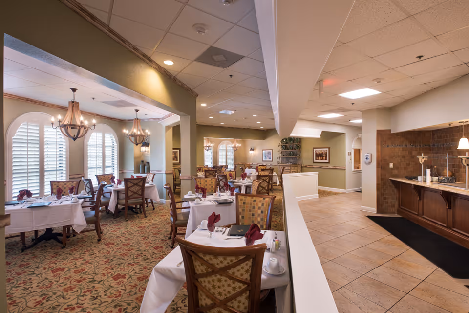 Elegantly set dining room with multiple tables, chandeliers, patterned carpet, and shuttered windows.