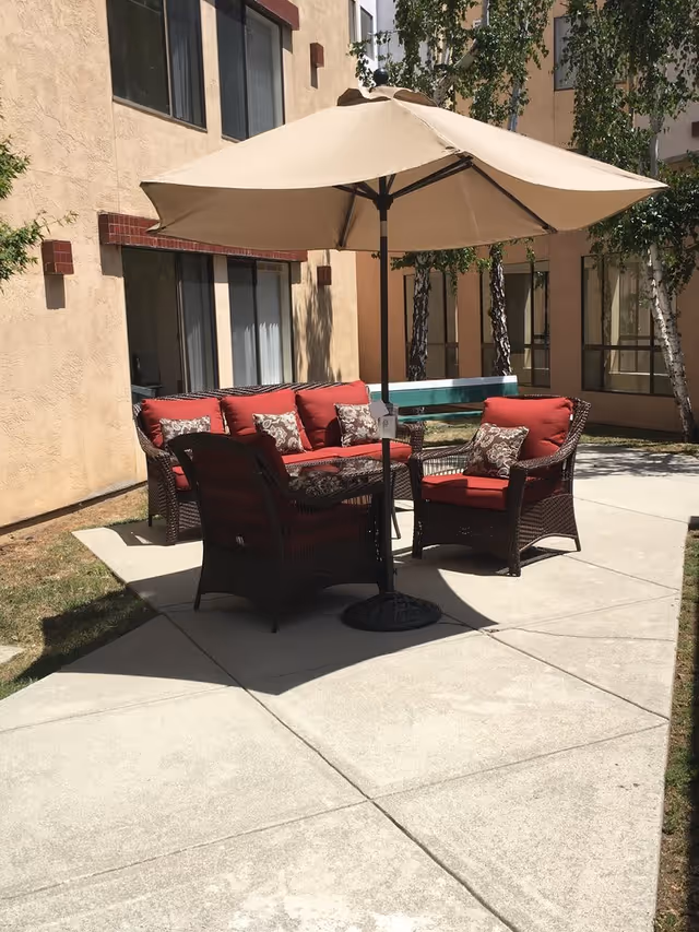 Outdoor patio area with a beige umbrella shading a set of dark wicker furniture with red cushions and patterned throw pillows, situated on a concrete surface next to a building with beige walls and windows. Trees and grass are visible in the background.